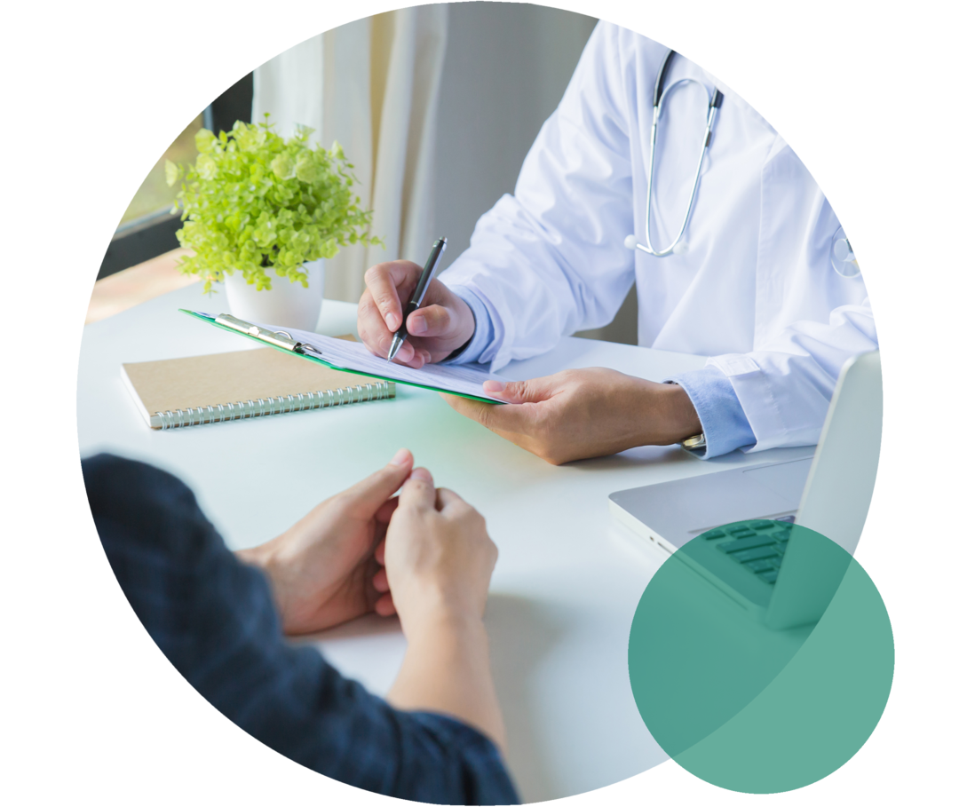 Zwei Personen sitzen sich an einem Tisch gegenüber. Eine Person mit weißem Kittel hält ein Klemmbrett und notiert etwas. Die zweite Person wartet mit gefalteten Händen.