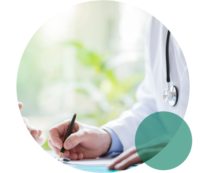 Person in weißem Kittel hält einen Stift und notiert etwas. Gegenüber sind die gefalteten Hände einer weiteren Person zu sehen.