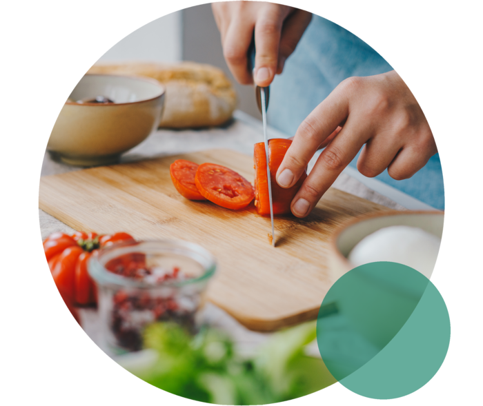 Küchenbrett auf einer Arbeitsplatte. Zwei Hände schneiden eine Tomate mit einem Messer.