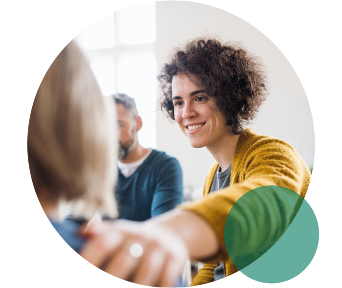 Zwei Frauen sitzen nebeneinander. Eine Frau legt lächelnd die Hand auf die Schulter der anderen Frau.