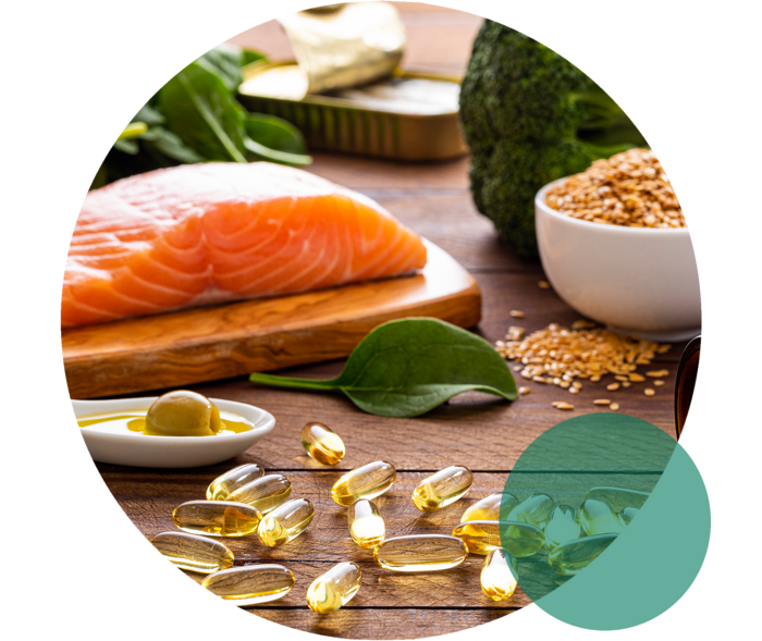 Auf einer Fläche liegen Lachs, Blattspinat, Brokkoli, ein Löffel mit Olivenöl sowie gelbliche Kapseln.