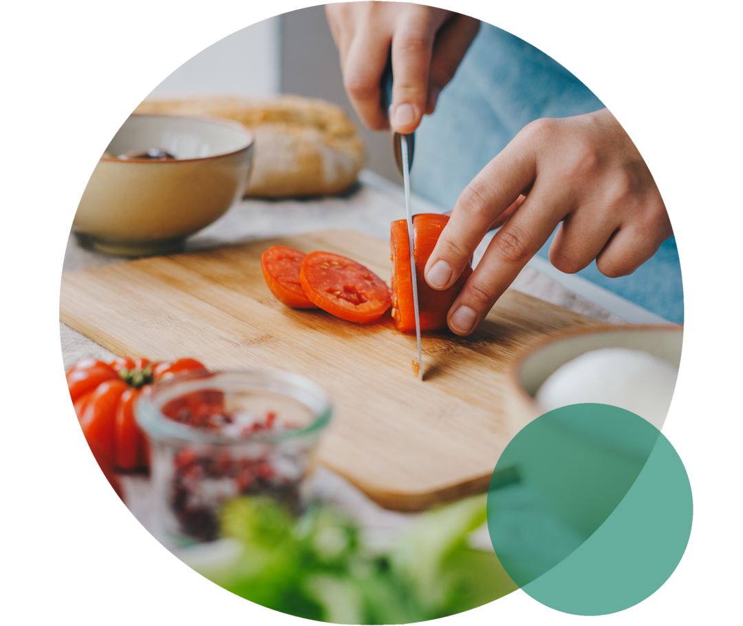 Küchenbrett auf einer Arbeitsplatte. Zwei Hände schneiden eine Tomate mit einem Messer.