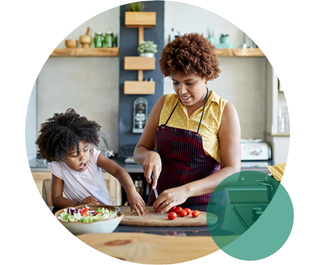 Eine Frau mittleren Alters mit Küchenschürze. Sie schneidet lächelnd Tomaten. Neben ihr steht ein Kind, das ihr zusieht.