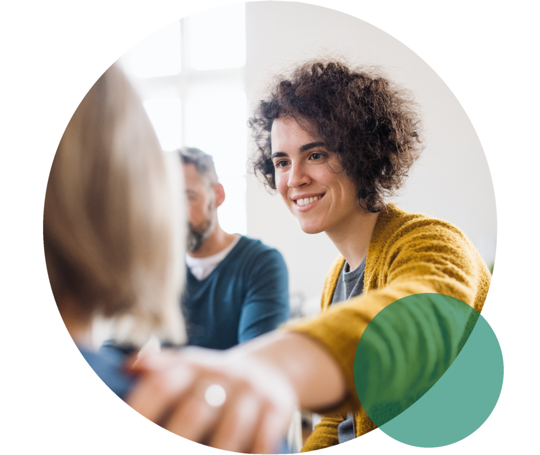 Zwei Frauen sitzen nebeneinander. Eine Frau legt lächelnd die Hand auf die Schulter der anderen Frau.