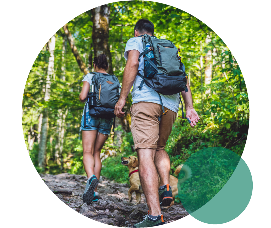 Eine junge Frau und ein junger Mann in sommerlicher Wanderbekleidung. Sie gehen bergauf auf einem Wanderweg, der von Bäumen umsäumt ist.