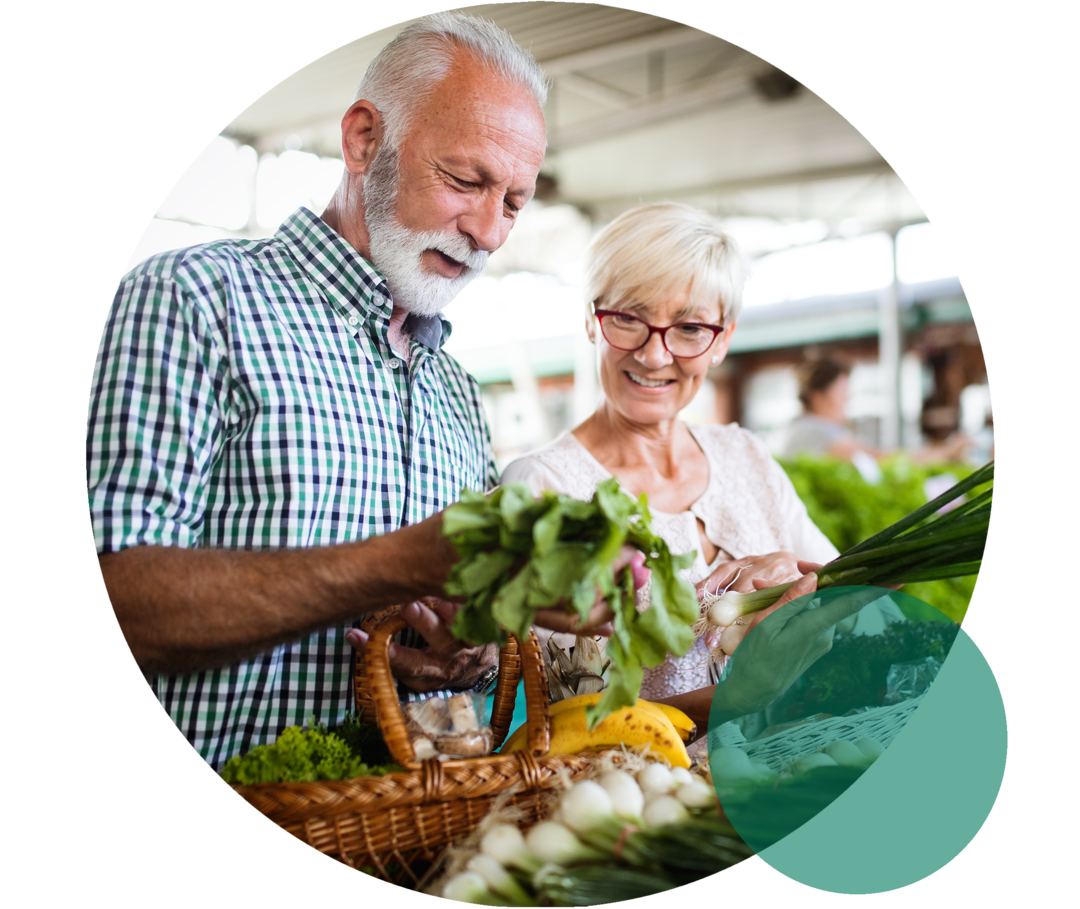 Ein älteres Ehepaar steht lächelnd an einem Marktstand mit Gemüse. Der Mann hält einen Salatkopf in der Hand, den beide betrachten.