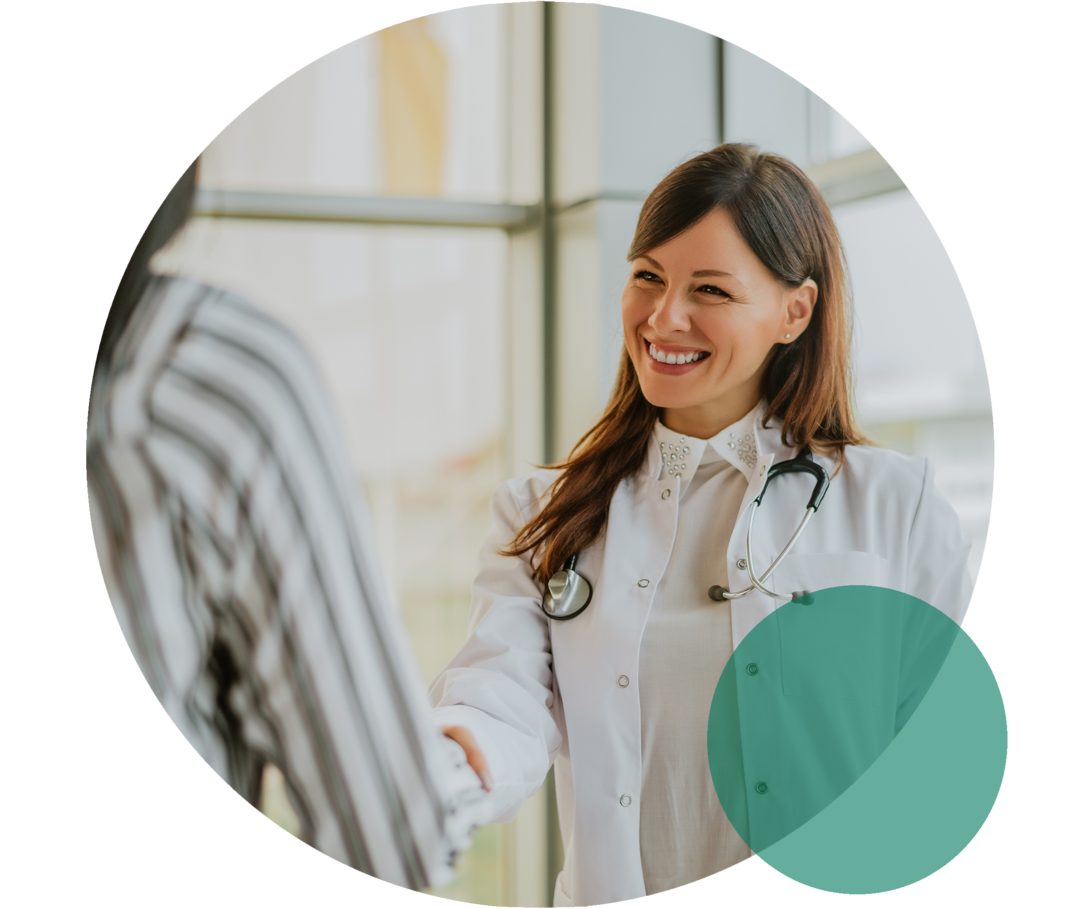 Junge Ärztin mit weißem Kittel reicht einer Person die Hand und lächelt sie an.