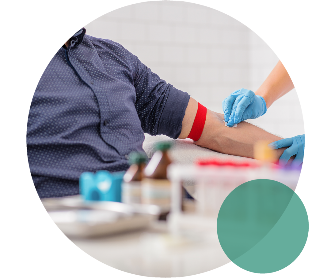 Ein Mann sitzt auf einem Stuhl. Neben ihm steht eine Person in weißem Kittel. Sie trägt Einweghandschuhe und streift mit einem Alkoholtupfer über die linke Armbeuge des Manns.