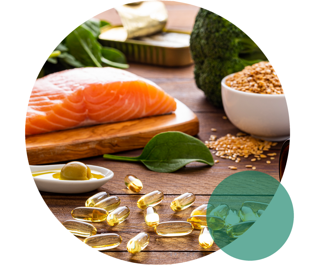 Auf einer Fläche liegen Lachs, Blattspinat, Brokkoli, ein Löffel mit Olivenöl sowie gelbliche Kapseln.