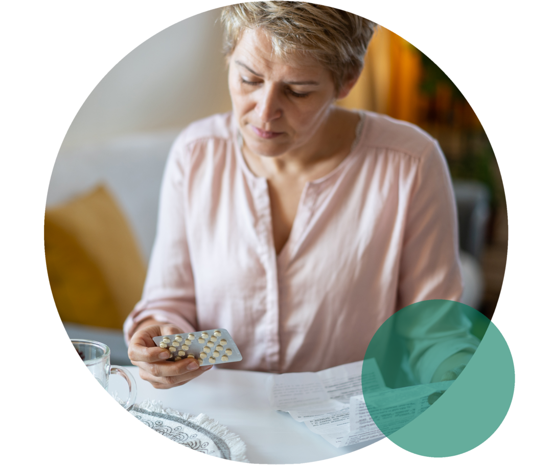 Eine Frau sitzt an einem Tisch. Sie hält ein Tablettenblister in der rechten und einen Beipackzettel in der linken Hand. 